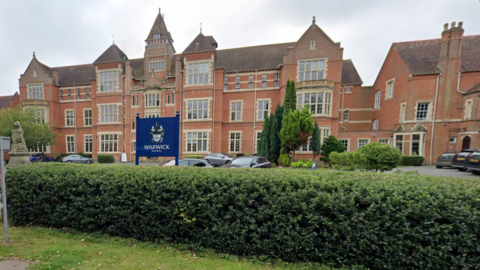 A Google street view image of red-brick Victorian school building on three levels, with a fourth level in a tower. A blue sigh in front of the building with a crest on it says Warwick School. A hedge is at the edge of the site and cars parked in front of it.