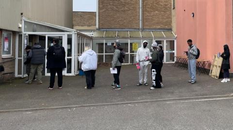 Ten people stand in a line at the door to a building. Groups of people in the line are spaced out.