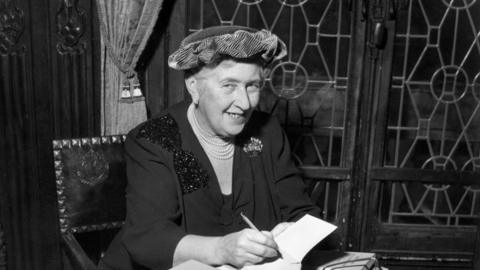 A black and white photo of Agatha Christie sitting on a chair as she signs a book. She is smiling at he camera and is wearing a hat, a black blouse and a pearl necklace.