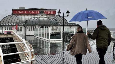 A couple with an umbrella walk along a wet Cromer pier under a grey sky