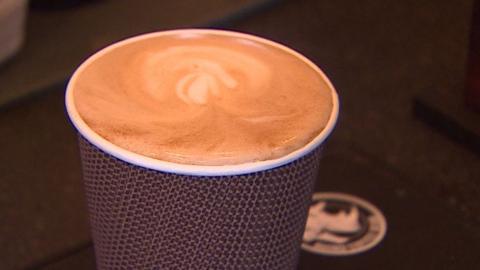 Brown takeaway coffee cup filled to the brim with coffee. The top froth has a caramel coloured swirl to it and a paler pattern drawn through it