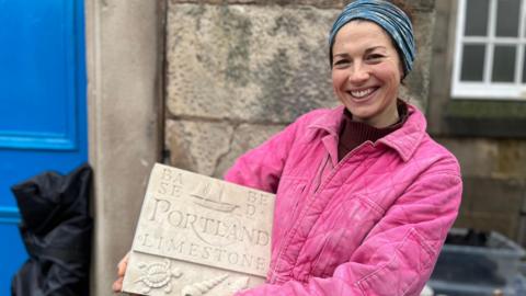 A smiling woman with her dark hair tied up, wearing pink overalls, holds a slab of carved limetone