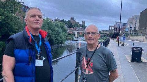 Tony Jones, left, and Gav are two middle-aged men. Toby is wearing a dark blue gilet and black t-shirt and Gav has spectacles and is wearing a grey t-shirt. Both have close-cropped hair and are wearing lanyards with an identity badge. They are standing on a street next to a railing with the river to one. Durham Cathedral and Castle can be seen in the far background.