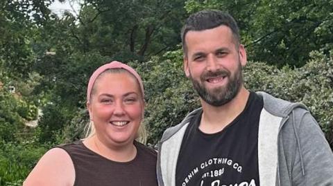 Sophie Cole, who has her blonde hair tied back by a pink headband. She is wearing a black vest and is smiling. She is standing next to her partner, who has short black hair, a beard and is wearing a black T-shirt and grey jumper. They are standing in front of trees.