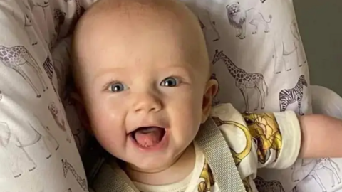 A baby sitting in a white baby seat with animals on it is smiling at the camera