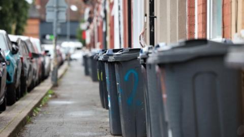 A picture of black bins lined up on a pavement. One of the bins has a blue '2' spray painted on it.