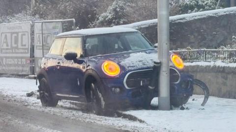 A blue mini crashed with orange lights on crashed into a bollard and lamppost in snow.