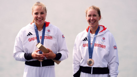 Two women dressed in white tracksuits with Great Britain across the chest in red lettering, while wearing bronze medals.