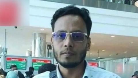 Mizanur Rahman is seen with glasses, a beard, and short black hair stands in an airport near  check-in counters, wearing a light-colored shirt.