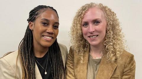 Two women sat together smiling at the camera. The woman on the left has braids that are half pulled up and she had a black t-shirt, gold necklace with a pendant and a cream blazer. The woman on the right has blonde curly hair, a beige t-shirt and a dark beige blazer