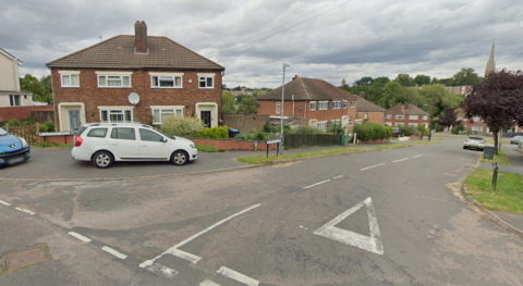 A road junction on a residential street lined with semi-detached houses. There is a white car parked on the pavement