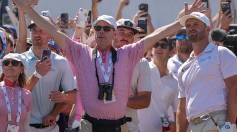 Wyndham Clark watches after hitting a shot while a marshal holds his arms high to signal to the crowd to keep quiet while he plays