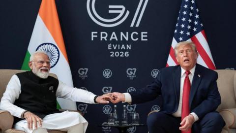 Modi, wearing white and black, holds the hand of Donald Trump, who is wearing a suit and red tie. They sit on chairs in front of an Indian and a US flag, with a 'G7 Evian 2026' sign in the background