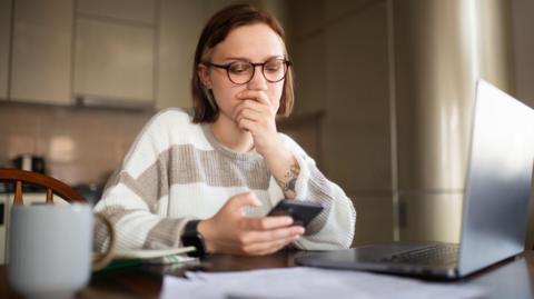 A young woman with round glasses and short brown hair wearing a striped white and beige long sleeved top and a smart watch is sat at a kitchen table holding her smartphone with her laptop open, papers in front of her, and tea by her side. She appears to be struggling to find job opportunities online and her hand is over her mouth.