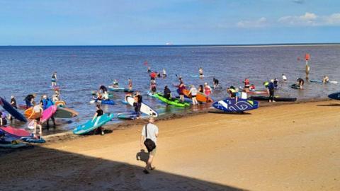 A group of paddleboarders on the sand walking into the sea with their boards that are all different colours. Another man wearing shorts and a white top and black bag walks by.
