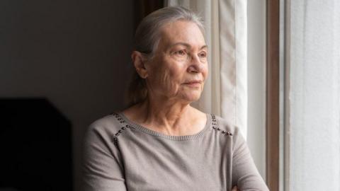 An older woman with long silver hair looks out a bedroom window. She wears a pale grey top with silver beads around the shoulders.