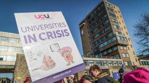 Protesters outside a university building hold a UCU placard reading “Universities in Crisis” during a daytime demonstration.