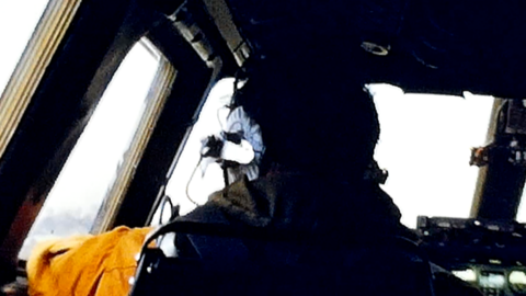 A low-quality image of the back of a pilot's head and body. The pilot is sitting in a cockpit wearing a helmet and headphone set. He is looking out of the window to his left.