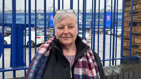 a woman with grey hair and glasses on her head stood in front of blue railings at a football stadium. She is wearing a black coat and a multi coloured scarf with blue, purple and pink sections