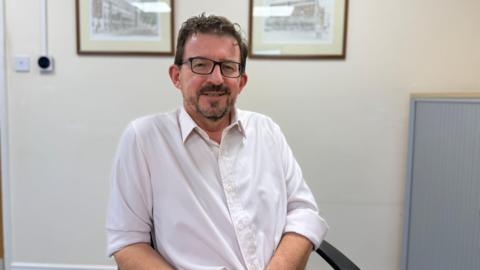 A picture of Owen Cole who is a middle aged man with a short dark beard. He has short dark hair which is slightly curly on top. He is wearing a white shirt with his sleves rolled up. There is artwork behind his head, one on the right hand side and one on the left hand side which are out of focus.