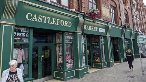 A market building with a green and gold front displays large “CASTLEFORD MARKET” lettering. People walk past shop windows showing sweets, pet supplies and clothing.