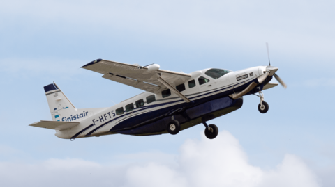 A Cessna Grand Caravan aircraft is flying against a backdrop of blue sky and some clouds. It has 'Finistair' written on it and is white and blue.