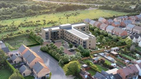 An artist's impression image of the new care home building, a large T-shaped structure surrounded by homes and greenery on one side, taken from a drone.