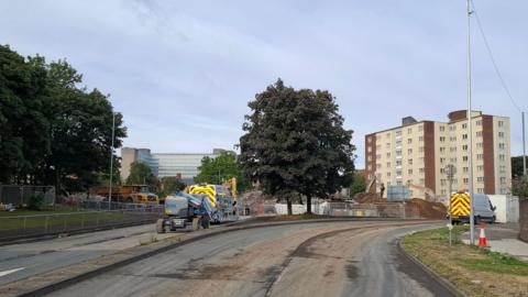 Park Lane in Gateshead, where demolition work on the Gateshead Flyover has been carried out. The road is closed and there are two vans and a number of heavy plant machinery, including a digger. There is metal fencing around the junction, with mounds of rubble and dirt around the area being worked on. In the distance there are two blocks of flats and another large building.