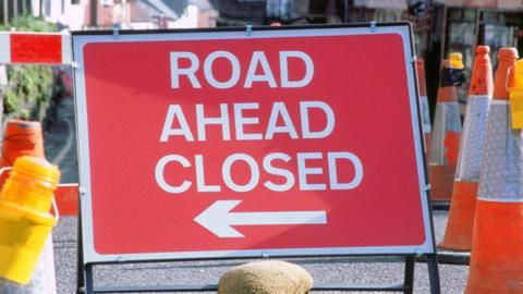 A generic shot of a red road sign reading "road ahead closed" in white lettering. It includes an arrow pointing to the left. It is surrounded by several orange traffic cones. 