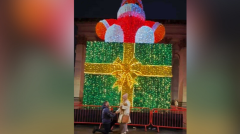 A man is down on one knee in front of a Christmas light display in Manchester's St Peter's Square. The lights show a big green present wrapped with a bow and a big orange Santa Claus positioned on top. The man has brown hair and wears a raincoat the woman, blonde has long hair, wears a pink wooly hat and a white coat.