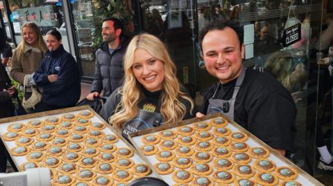 Ella Henderson wearing a black hoodie and black apron next to her friend Matty, they are both holding up large baking trays with round biscuits on. In front of a shop window with people standing around behind.