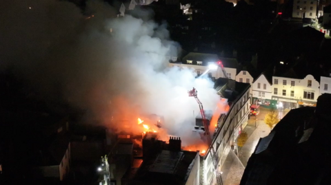 A drone shot of smoke coming from a building's roof which is on fire. There are two aerial platforms above the fire.