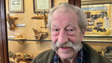 Fred Clarke, 82, has grey hair swept back over his head and a large handlebar moustache he is wearing a brown fleece with checked shirt underneath. Behind him is a display case containing some of his models including a traction engine, spitfire aircraft, and more, all made of wood and brass and highly detailed.