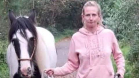 Leanne Williams wearing a pink hoodie. She is holding a rope guiding a brown and white horse beside her. They are in a wooded area. 