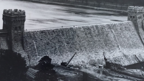 A black and white image of Derwent Dam, a large brick wall with two towers at either end. Cranes and trees can be seen at the bottom of the dam and there is a body of water to the left of it.
