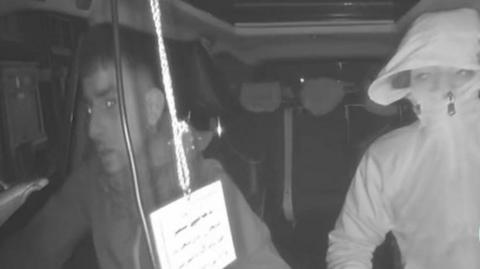 A grey screenshot image of CCTV footage, showing two people sat inside a car. The driver is looking forward, wearing a hooded jacket, with the passenger seen with a jacket zipped high over his face. A lanyard is hanging from the rearview mirror with writing on