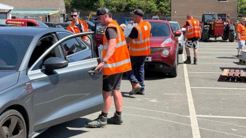 A number of staff wearing high-vis orange jackets provide bottled water to people sitting in cars. 