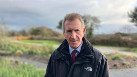 A man wearing a black North Face coat. He is wearing a blue shirt and red tie. He is looking directly at the camera. In the background is a garden. The sky is blue with clouds. 