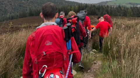 A group of mountain rescuers, viewed from behind, with back packs and rescue gear carry someone on a stretcher down off a very grassy hillside