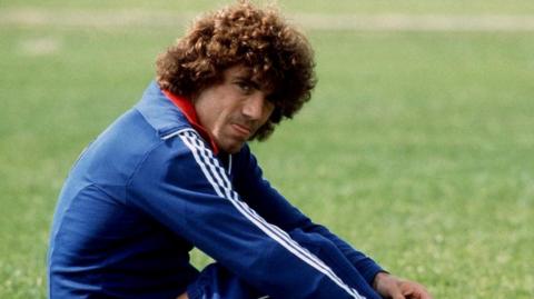 Kevin Keegan with 70s perm  in blue tracksuit with white stripes on the sleeves is sitting on the grass durign a training session with Hamburg SV