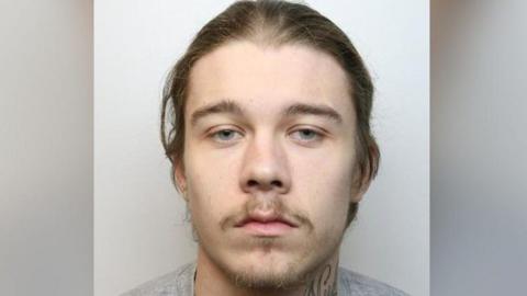 A mugshot of Ryan Malloy. He has long brown hair and a small amount of facial hair above his mouth and on his chin