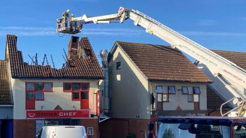 Another view of the scene of the fire. A cordon has been placed around the building where the fire took place. A fire engine can be seen to the right as an aerial ladder hovers near the building's roof that has been damaged.