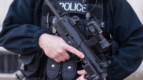 A close up of an armed Police Officer holding a gun with the words "police" written on his vest.