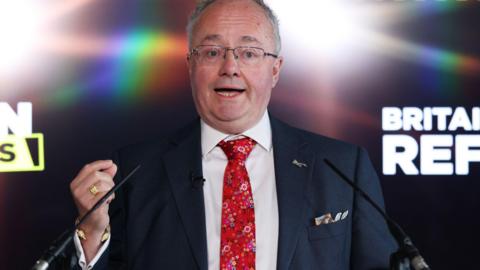 A man in a suit and tie in front a screen saying "Britain is Lawless"