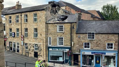A view of a street of tall, terraced, sandstone buildings. The two to the right are shops, with one of them, Bobs Barbers has a gaping hole in the tiled roof due to a collapsed chimney. Some rubble is strewn on the ground in front of the building and the street is taped off. 