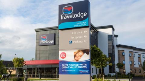 The outside of a Travelodge hotel with a nearby sign promising customers will "sleep soundly with great value rooms" beneath a picture of a guest smiling in bed.