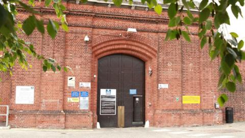 The large brown entrance door to HMP Norwich, which is in the middle of tall and wide brick walls. Branches of a leafy tree appear in the foreground of the image.