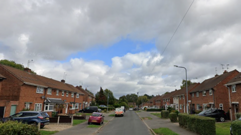 A residential street lined with brick houses, parked cars, and small front gardens beneath a cloudy sky.