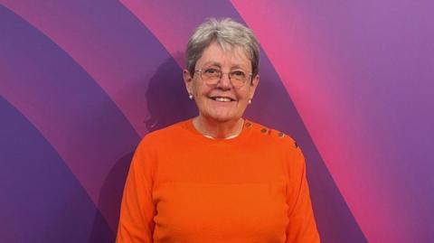 Janet McConkey, a woman wearing glasses and an orange jumper is standing against a prurple background.