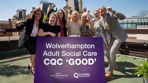 Seven people, six women and one man are standing on what looks to be a rooftop area of a building, around a large purple sign that reads "wolverhampton adult social care CQC good". They are cheering with their fists in the air in celebration